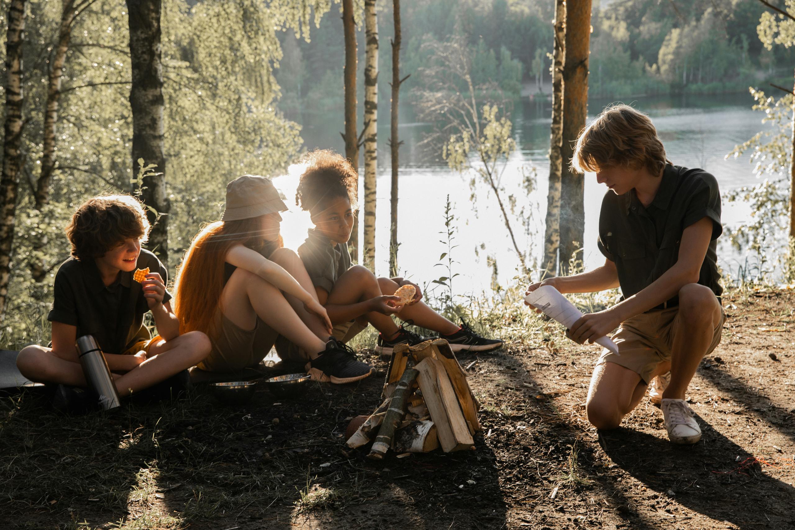 Children enjoying a camping trip by a lake, preparing for a campfire, surrounded by nature at sunset.