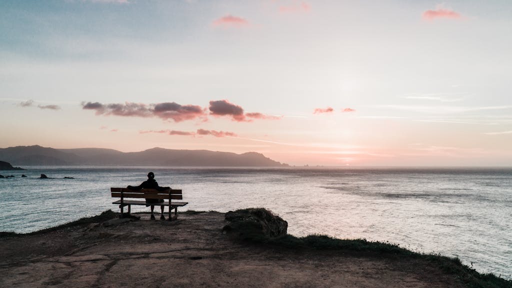 A peaceful sunset view of a person sitting on a bench by the ocean, capturing tranquility.